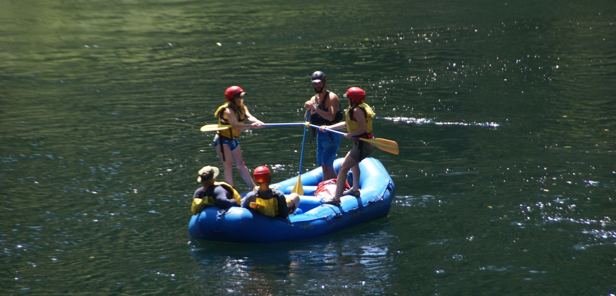 A group of five river rafters in a blue raft balance an oar in their boat along the Sandy River.