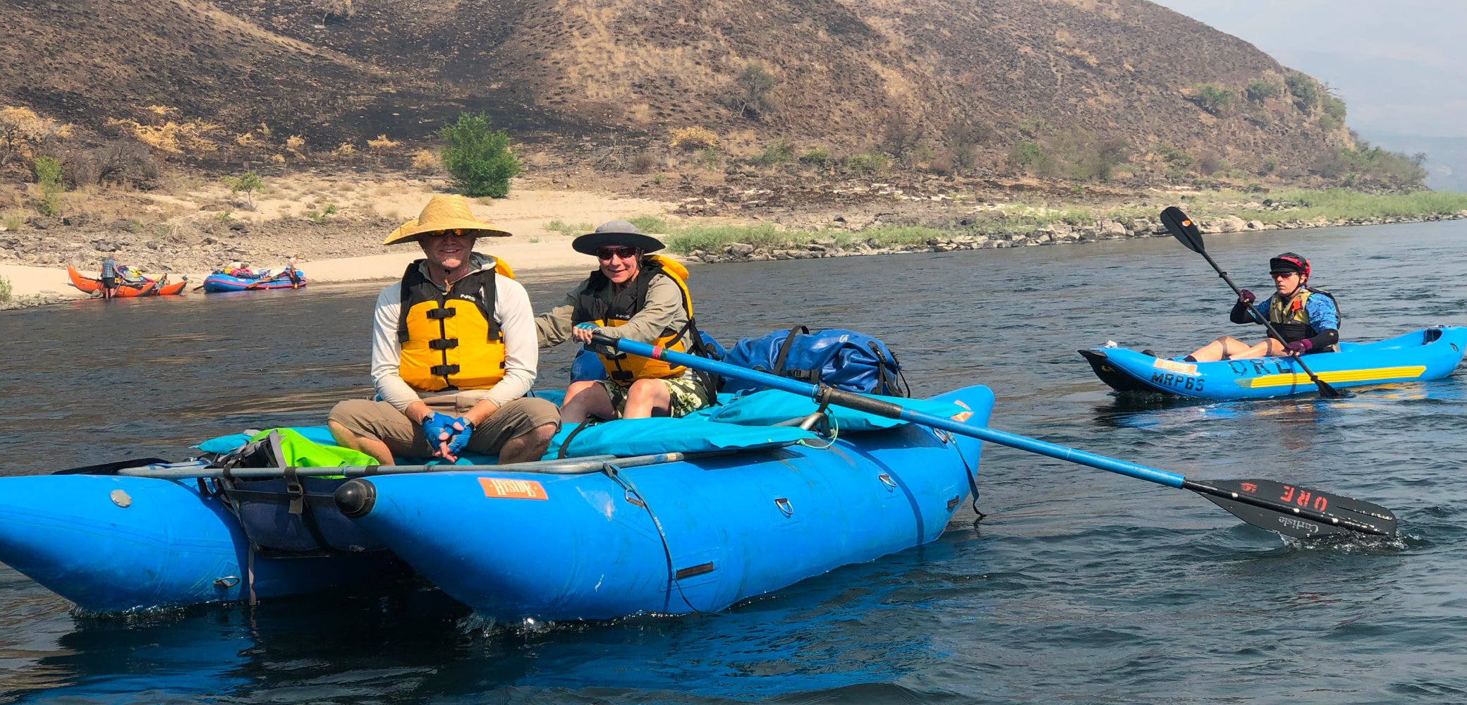 Two people in life vests ride atop a blue cataraft float in the river while another floats in a blue inflatable kayak.