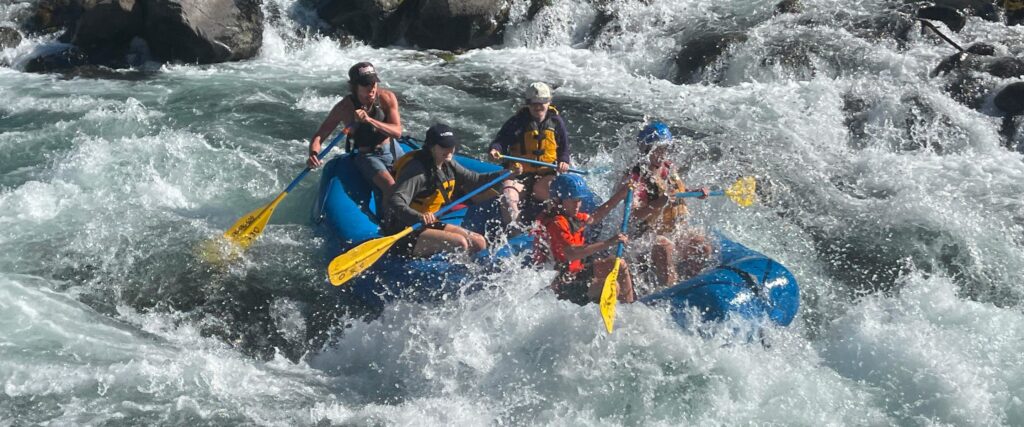 A group of white water rafters on a blue raft paddle through a section of river rapids on the Clackamas River in Oregon.