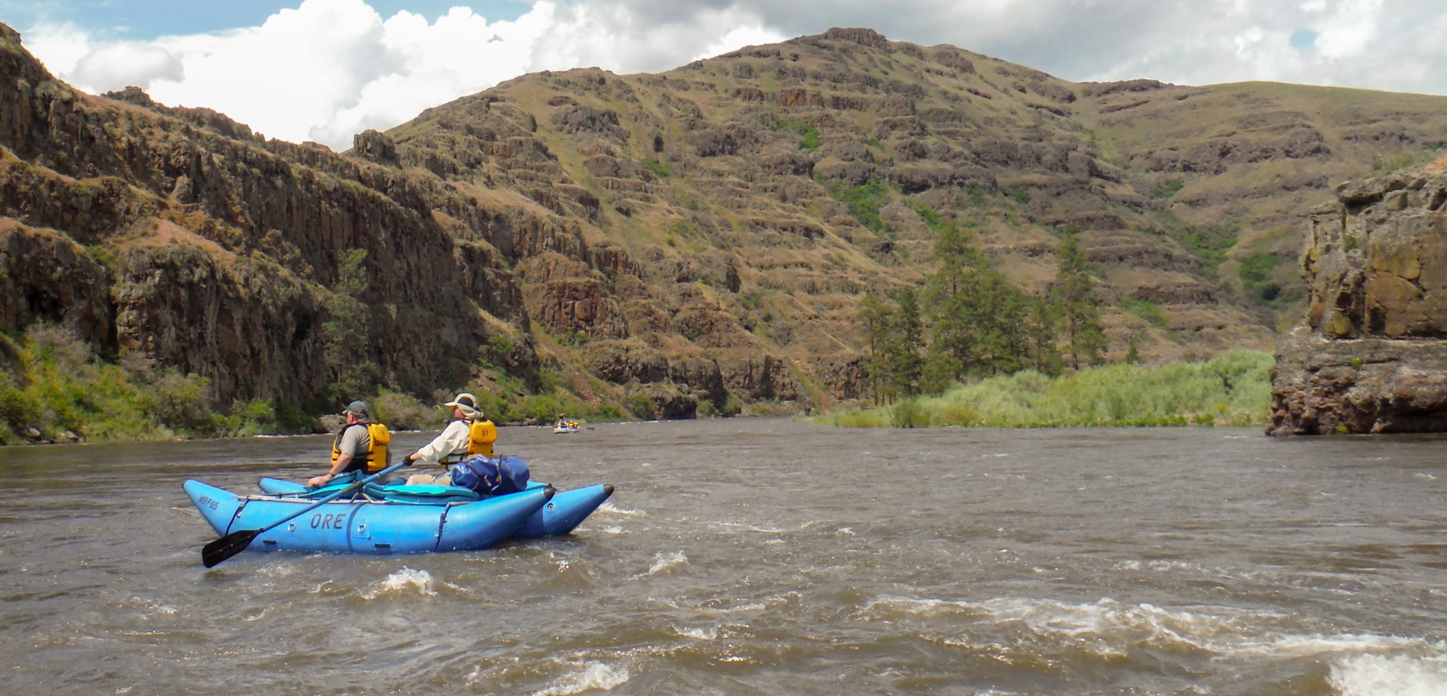 Two people in life vests ride atop a blue cataraft while navigating a calm river in Oregon.
