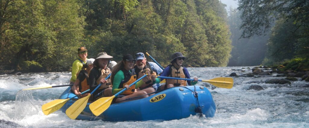 A group of smiling white water rafters on a blue raft paddle through a section of river rapids on the McKenzie River in Oregon.