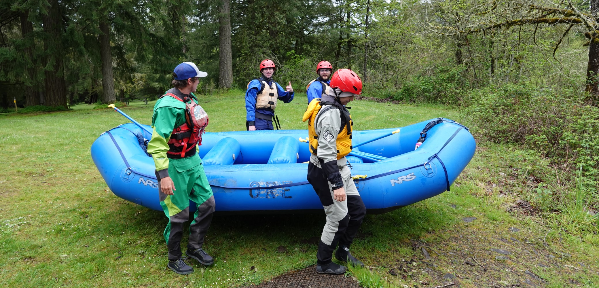 A group of white water rafters carry a blue raft toward the river while in full gear.