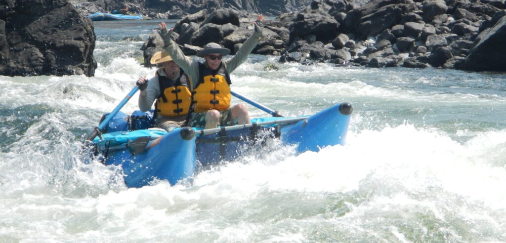 Two people in life vests ride atop a blue cataraft while navigating white water rapids in Oregon.