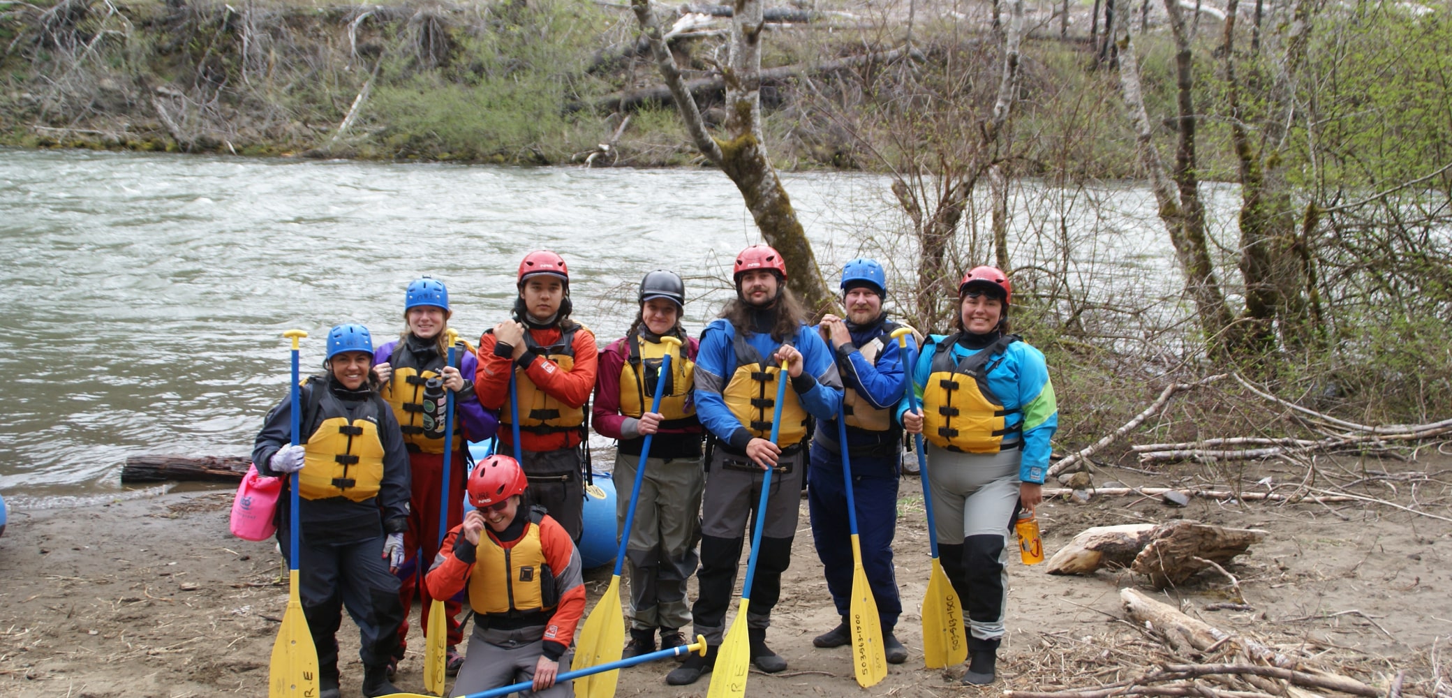 A group of 8 river guide trainees standing with oars, helmets, and life jackets along the river bank.