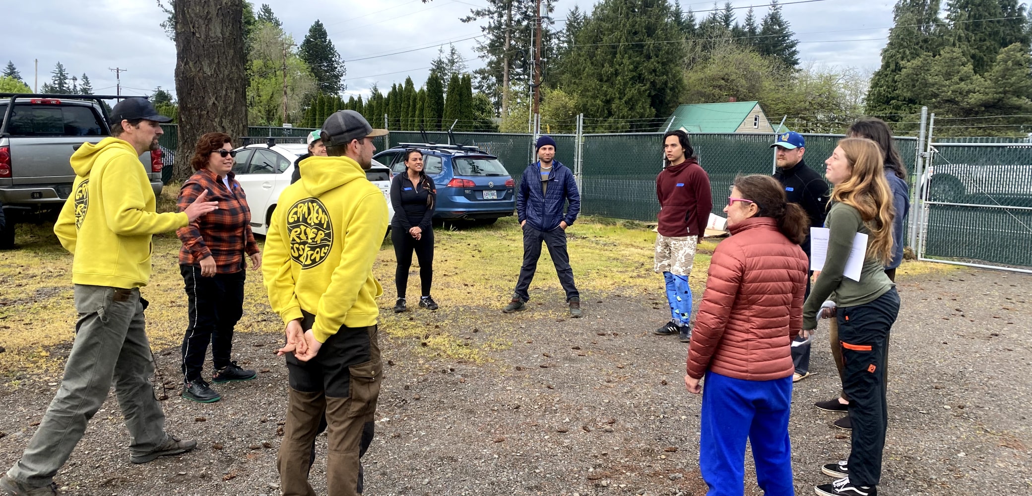 A group of 11 river guides stand in a circle for a training and safety meeting in a parking lot.